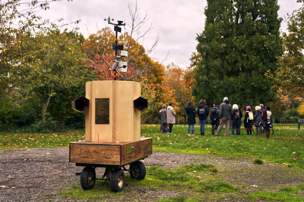 Future Machine in the memorial gardens in Finsbury Park with the procession in the background and trees behind