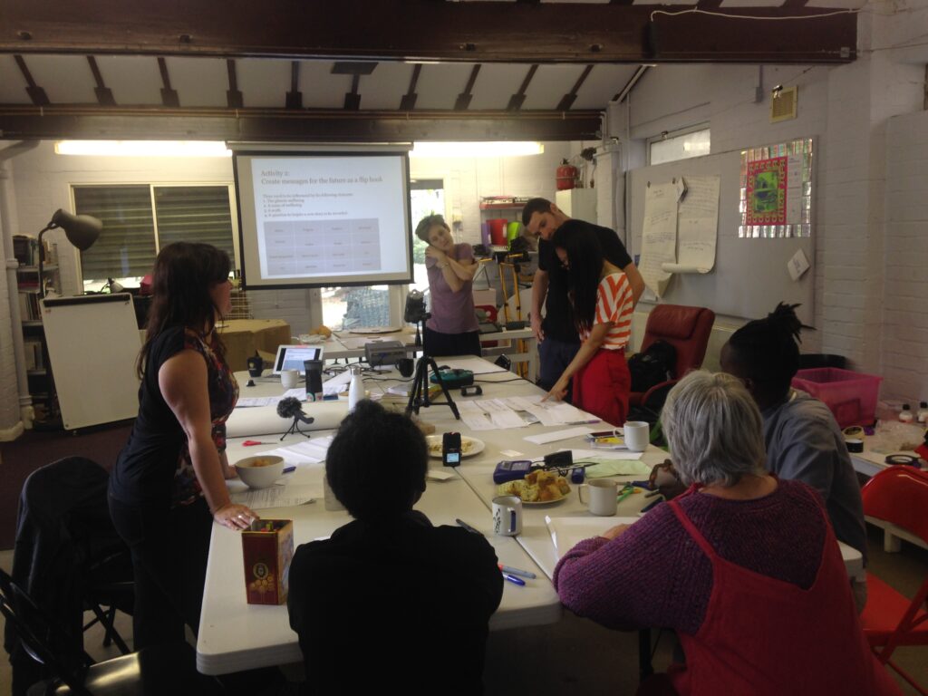 Rachel and participants standing around a table with materials on it. With a projection in the background titled 'Building a future flip book'